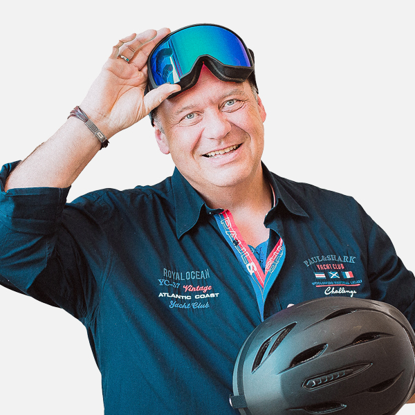 Frank Frohme, mit einem breiten Lächeln, trägt ein dunkelblaues Hemd, hebt eine Skibrille an und hält einen schwarzen Helm in der Hand, vor weißem Hintergrund.