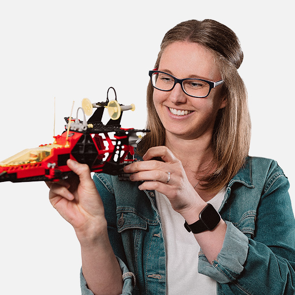 Imke Backhaus hält ein detailreiches LEGO-Raumschiff in den Händen und betrachtet es mit einem Lächeln. Sie trägt eine Jeansjacke über einem weißen Shirt und eine Smartwatch am Handgelenk.