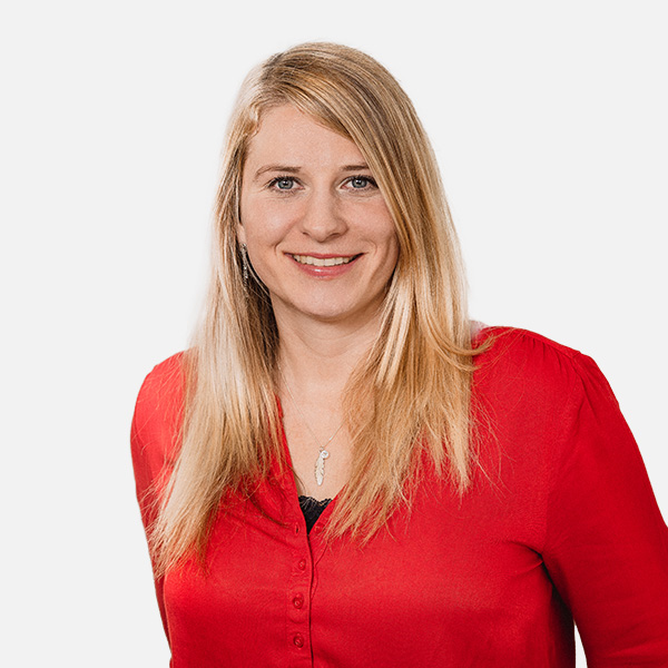 Jana Grube mit langen, blonden Haaren, lächelt freundlich in die Kamera. Sie trägt eine rote Bluse und eine silberne Kette mit Anhänger. Der Hintergrund ist weiß.