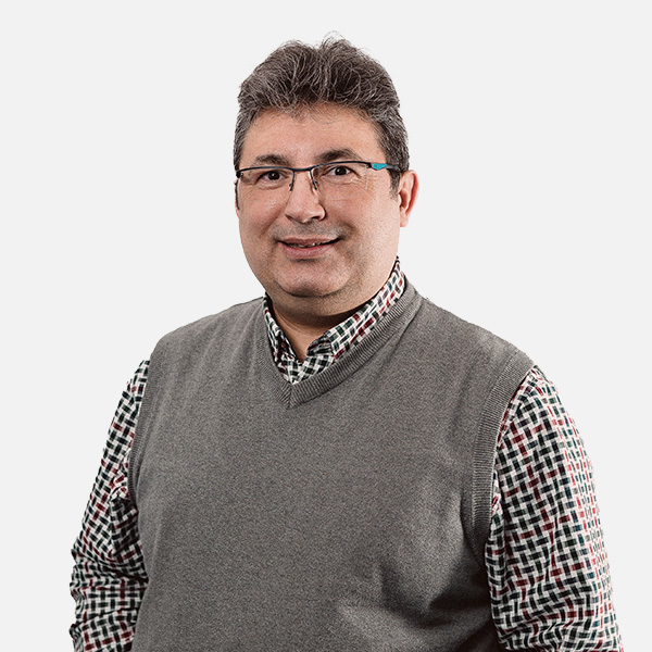 Kaya Küçük mit Brille, kariertem Hemd und grauer Strickweste, lächelnd vor weißem Hintergrund.