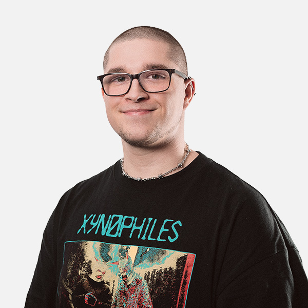 Leeon Sauer trägt ein schwarzes T-Shirt mit buntem Aufdruck und eine silberne Kette, während er freundlich in die Kamera lächelt vor weißem Hintergrund.