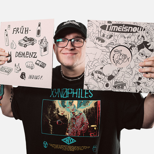Leeon Sauer trägt ein schwarzes T-Shirt mit buntem Aufdruck und eine schwarze Cap, während er zwei Schallplatten-Cover mit kreativem Design in die Kamera hält und dabei lächelt.