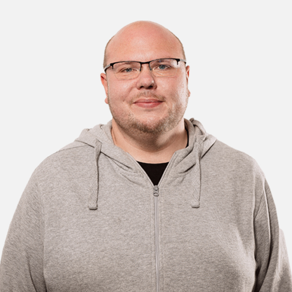 Marc Roba mit freundlichem Ausdruck, trägt eine graue Kapuzenjacke und eine schwarze Brille, vor weißem Hintergrund.