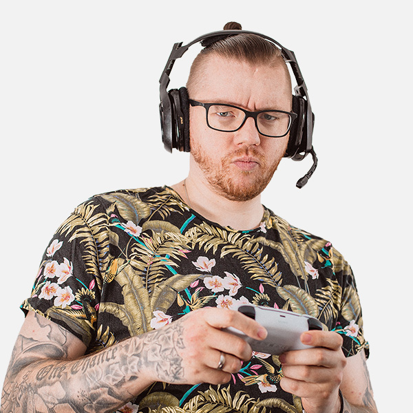 Mike van Doorn mit Headset und Brille, trägt ein bunt gemustertes T-Shirt und hält konzentriert einen Gaming-Controller. Seine tätowierten Arme sind sichtbar. Weißer Hintergrund.