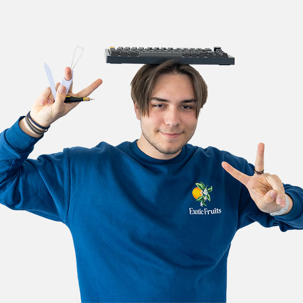 Tobias Hofmann mit spielerischer Pose, balanciert eine Tastatur auf dem Kopf, hält Werkzeuge in der Hand und trägt einen blauen „Exotic Fruits“-Pullover.