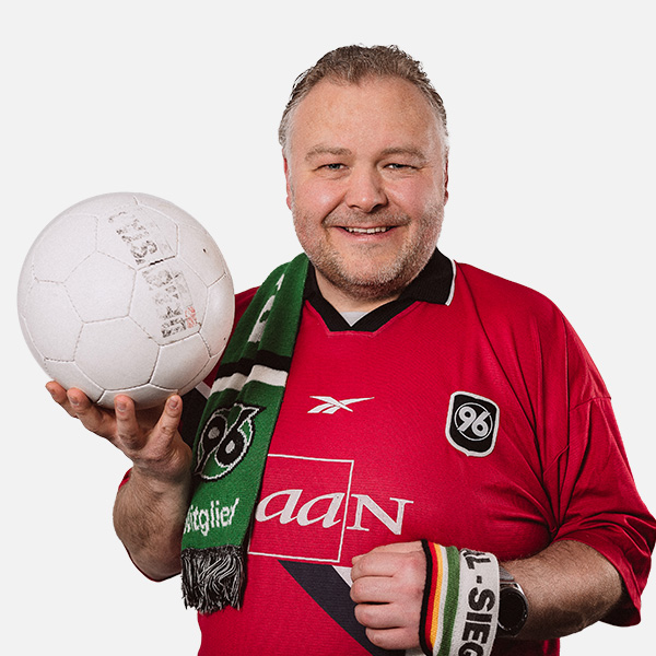 Peter Embscher, Geschäftsführer, in einem Hannover 96 Trikot mit Schal und Fußball, lächelnd vor neutralem Hintergrund.