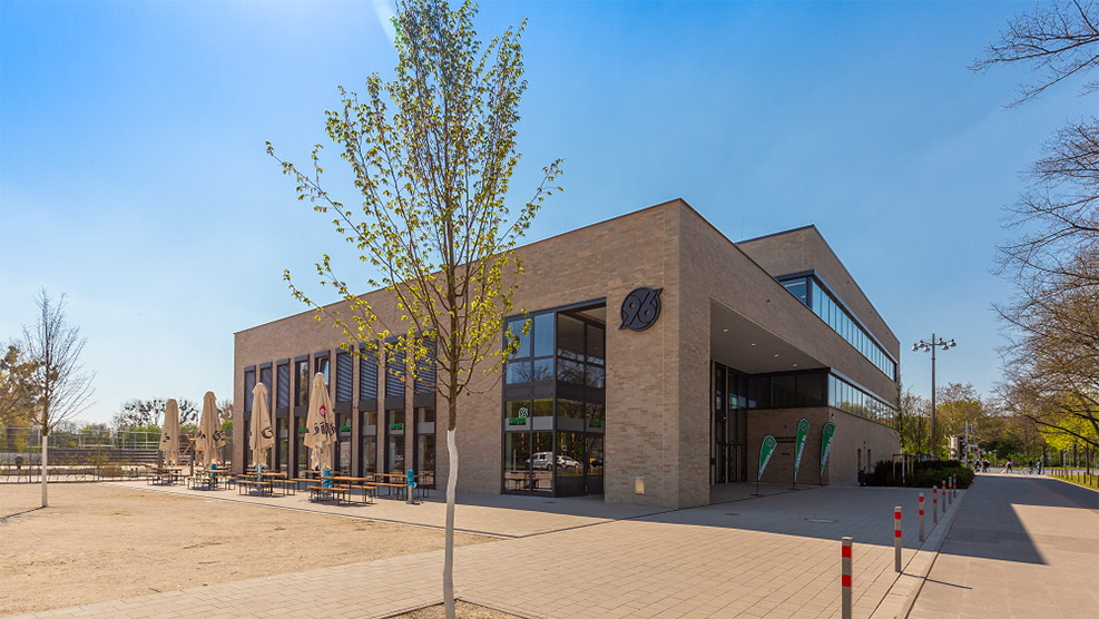 Das moderne Vereinszentrum von Hannover 96 mit Außenbereich, Sitzgelegenheiten und Hannover-96-Logo an der Fassade.