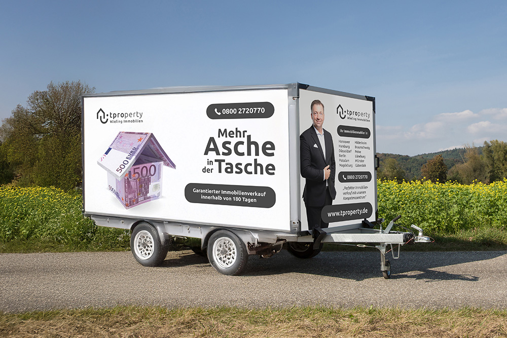 Werbeanhänger von tproperty mit Immobilienverkaufs-Werbung auf einer Landstraße, inklusive Slogan 'Mehr Asche in der Tasche' und Kontaktinformationen.