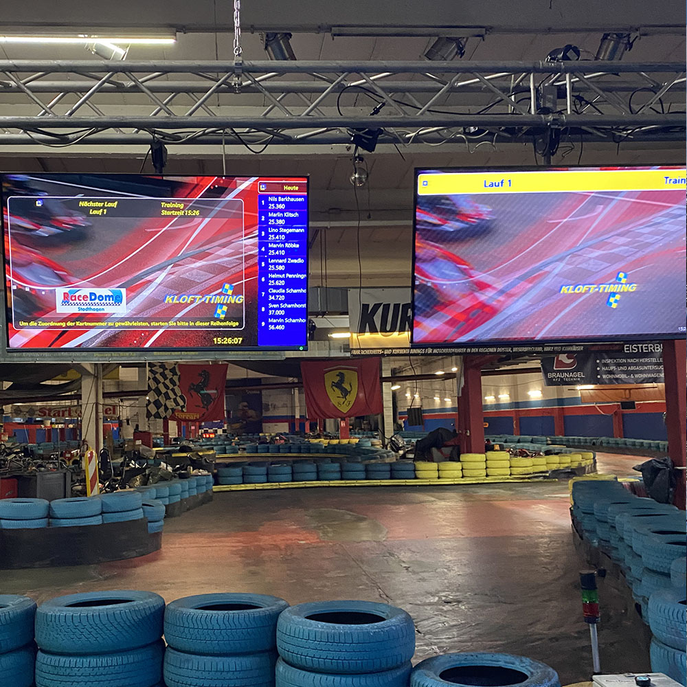 Teamevent auf der Kartbahn – Übersicht der Rundenzeiten auf großen Bildschirmen in der Indoor-Rennhalle vor dem nächsten Lauf