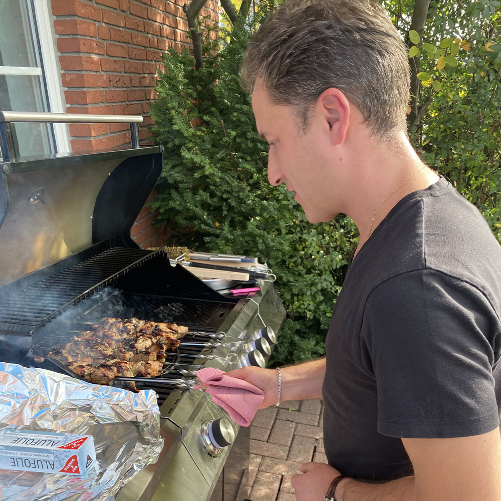 Teamevent mit Grillen in der Firma – Kollege am Gasgrill bereitet Fleischspieße vor, entspanntes Beisammensein bei sonnigem Wetter