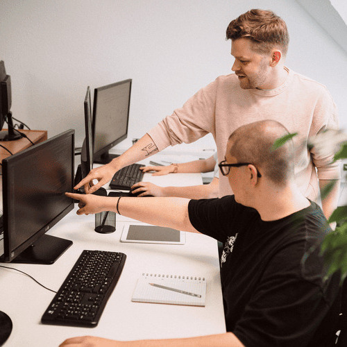 Zwei Webdesigner zeigen am Schreibtisch auf einen Bildschirm. Einer sitzt, der andere steht dahinter.