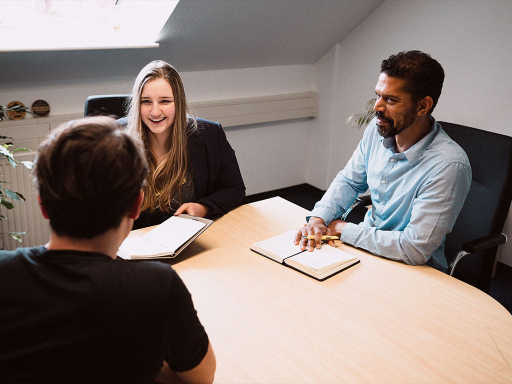 Drei Personen im Strategiegespräch für Online-Marketing am Besprechungstisch, mit Notizbüchern, in heller Büroumgebung.