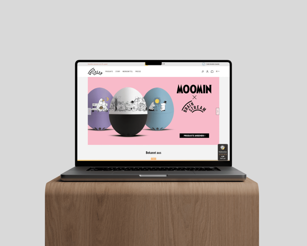 Aufgeklappter Laptop auf Holztisch zeigt Website von Brainstream mit MOOMIN-Produkten.