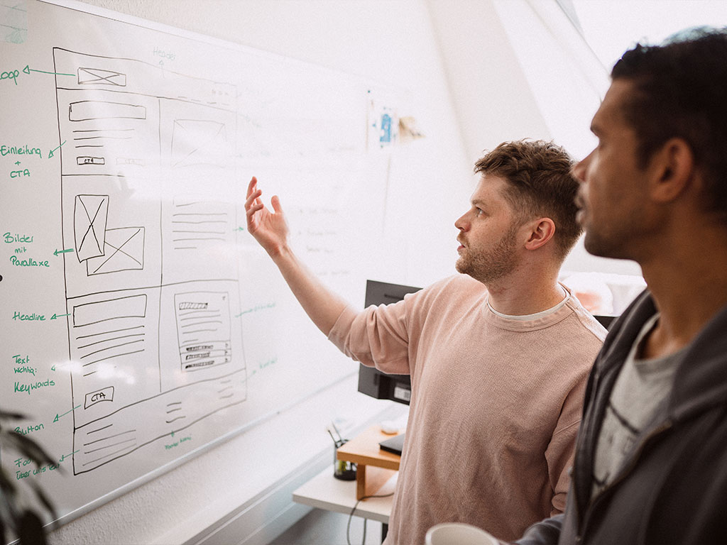 Zwei Kollegen stehen vor einem Whiteboard mit Website-Wireframes und diskutieren Struktur, Inhalte und Call-to-Action-Elemente.