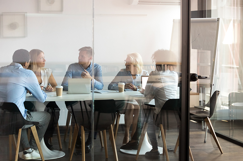 Fünf Personen sitzen an einem Tisch in einem verglasten Besprechungsraum und unterhalten sich, mit Laptops, Kaffeebechern und Flipchart im Hintergrund.