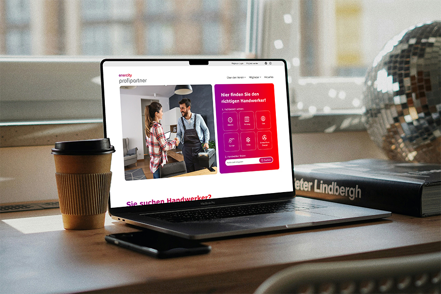 Laptop auf einem Schreibtisch mit geöffneter Website, daneben Kaffeebecher, Smartphone und Buch vor einem Fenster.