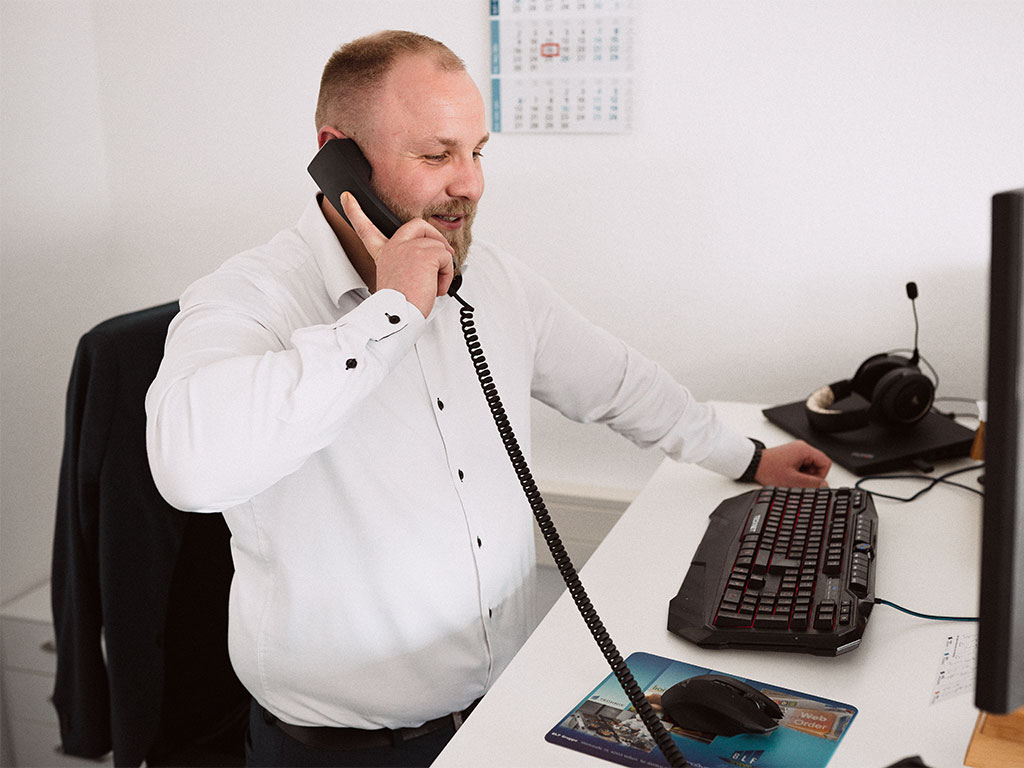 Mann mit kurzen Haaren in weißem Hemd telefoniert im Stehen am Schreibtisch – persönliche Beratung.